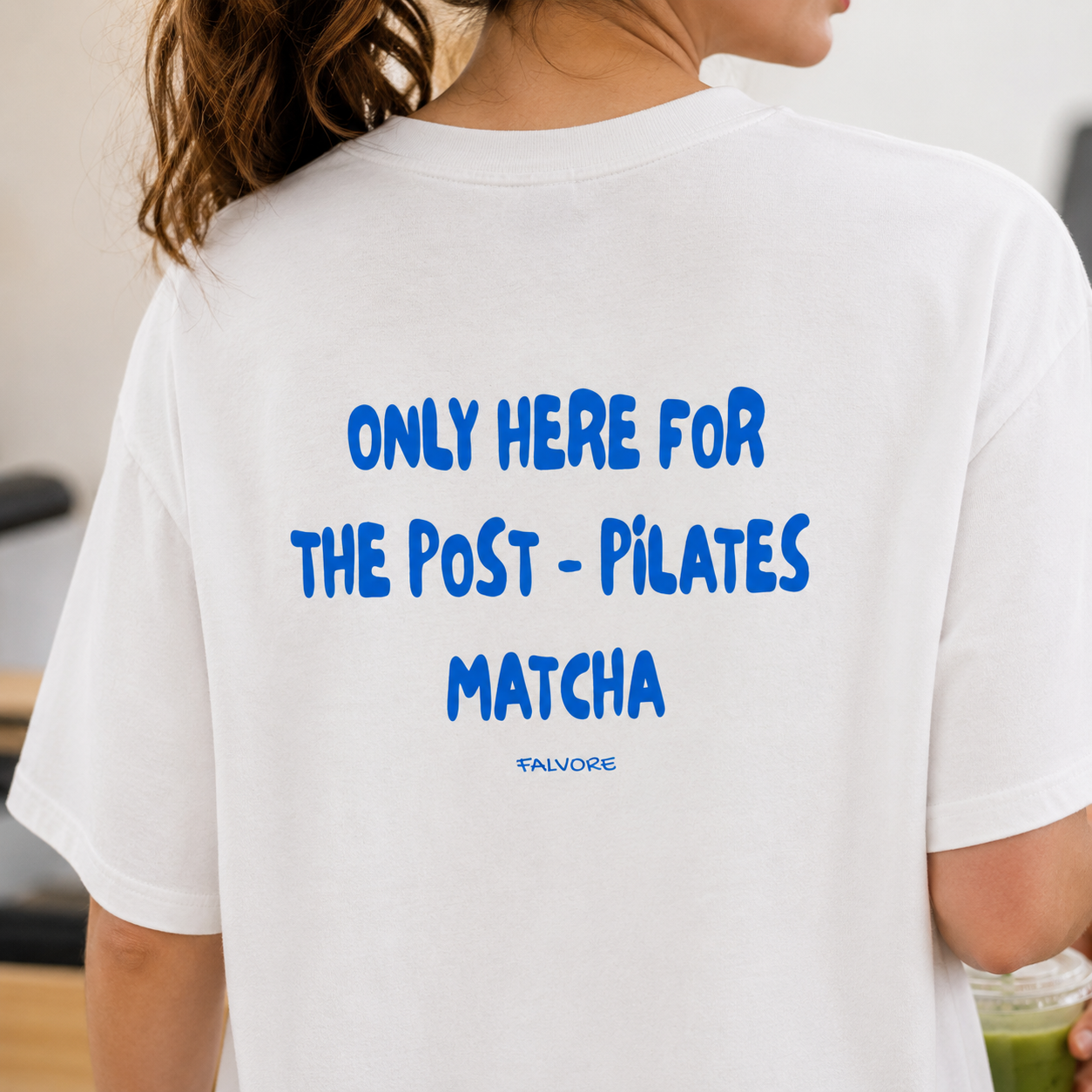 Person wearing a white t-shirt with blue text on the back, holding a green drink.