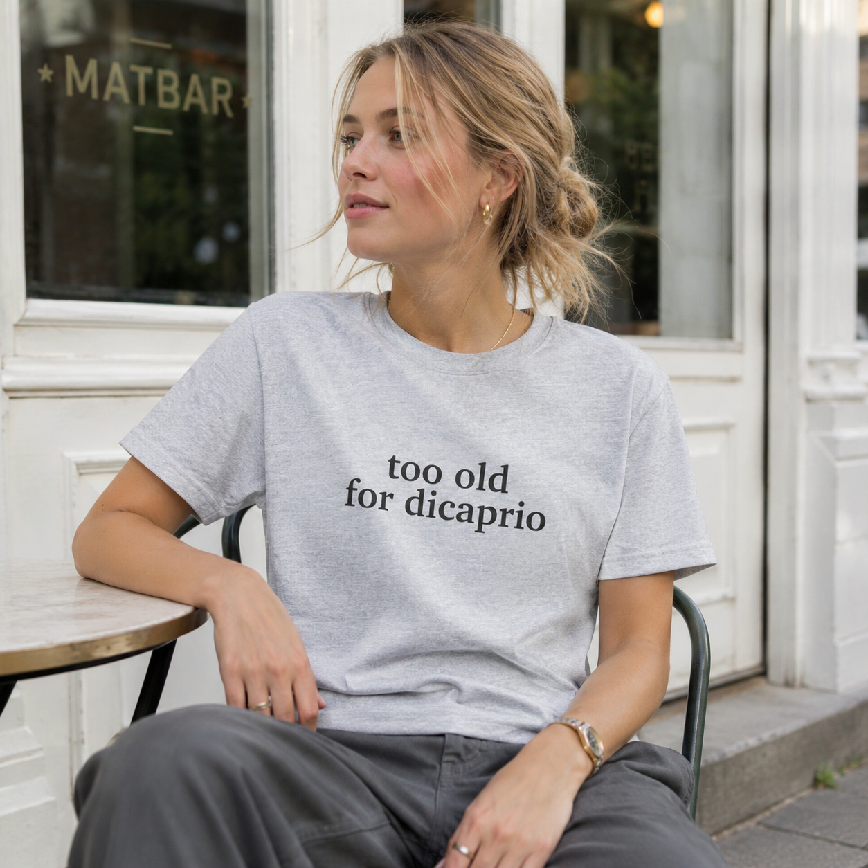 Woman wearing a t-shirt with 'too old for dicaprio' text sitting outside a building.